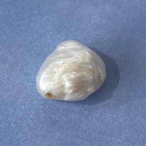 May include: A single, irregularly shaped, off-white pearl, possibly baroque, resting on a textured, light blue surface. The pearl has a lustrous, slightly translucent appearance, with subtle variations in color and texture.