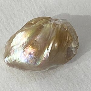May include: A large, irregularly shaped pearl with a lustrous, iridescent surface. The pearl displays a mix of colors, including gold, pink, and hints of blue and green. The surface has a textured appearance, with ridges and imperfections.