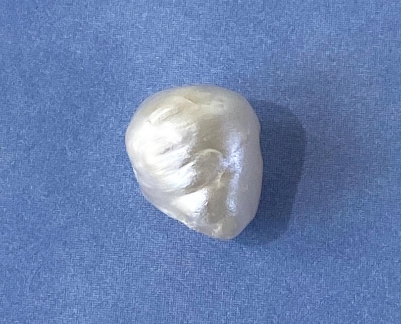 May include: A pearlescent, off-white, irregularly shaped object, possibly a pearl or a decorative element, rests against a textured blue background. The object has a smooth, reflective surface with subtle highlights and shadows.