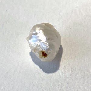 May include: A single, irregularly shaped, white pearl with a lustrous surface. The pearl has a slightly rough texture and a small, dark brown spot. The pearl is set against a white background, with a shadow cast to the lower left.
