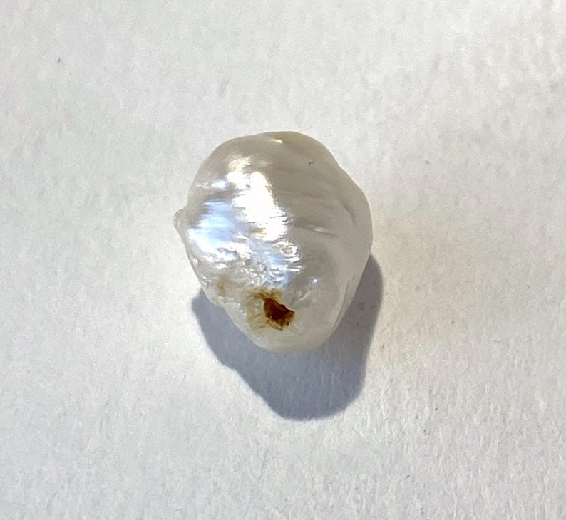 May include: A single, irregularly shaped, white pearl with a lustrous surface. The pearl has a slightly rough texture and a small, dark brown spot. The pearl is set against a white background, with a shadow cast to the lower left.