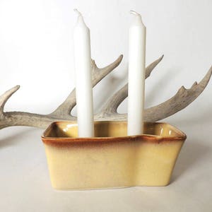 May include: A ceramic candle holder with a yellow glaze and brown accents. Two white candles are placed in the holder. The holder is sitting on top of a set of antlers.