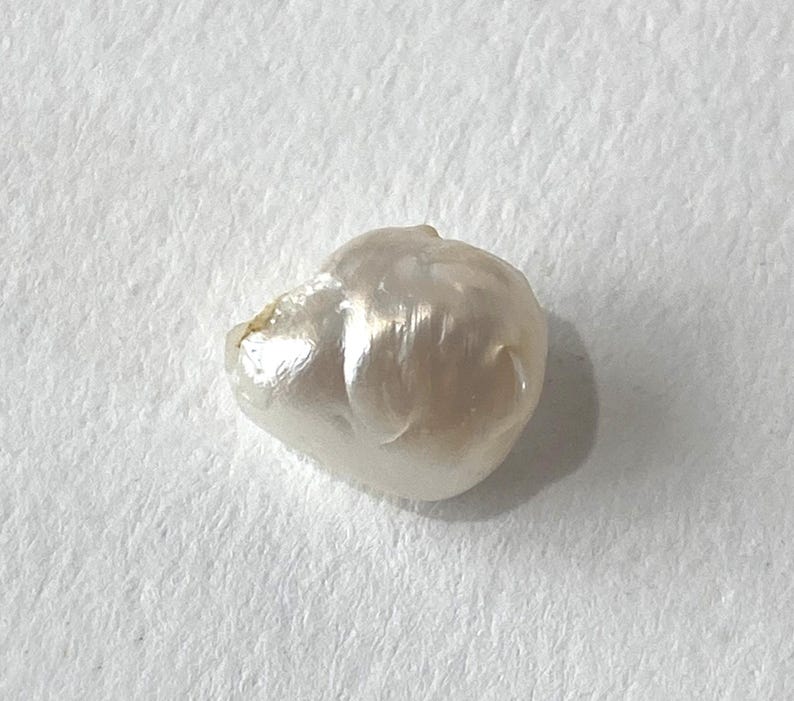 May include: A lustrous, irregularly shaped, off-white pearl. The surface reflects light, showcasing a natural, organic form. The pearl is set against a plain, textured white background.