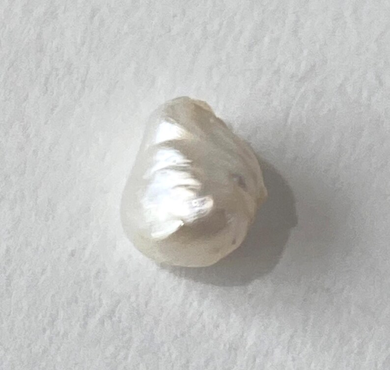 May include: A single, irregularly shaped, white pearl. The pearl has a smooth, lustrous surface with subtle variations in color and texture. The pearl is set against a plain, off-white background.