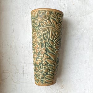 May include: A tall, ceramic vase with a detailed floral and leaf pattern in shades of green and brown. The vase has a textured surface and a slightly flared top, showcasing a nature-inspired design.