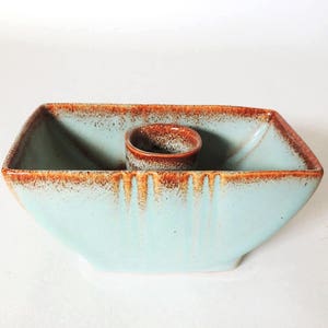 May include: A light blue ceramic candle holder with a brown glazed interior. The holder has a square base and a rectangular shape with a small round opening in the center for a candle.
