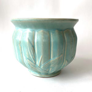 May include: A light blue ceramic planter with a ribbed design and leaf accents. The planter has a wide opening and a rounded base. The color is a soft, muted turquoise, and the overall shape is reminiscent of a classic flower pot.