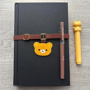 May include: A black notebook with a brown elastic band and a gold buckle. A yellow bear charm hangs from the band. A brown pen with the word "americano" is next to the notebook, along with a yellow bear pen.