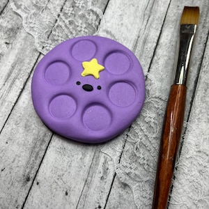 May include: Purple round paint palette with seven circular wells and a yellow star on top. The palette is on a white wooden surface with white lace.