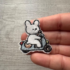 May include: Embroidered patch featuring a white bunny riding a light blue scooter. The bunny has black accents and is wearing a brown outfit. The scooter has black wheels and a red taillight. The patch has a white border.
