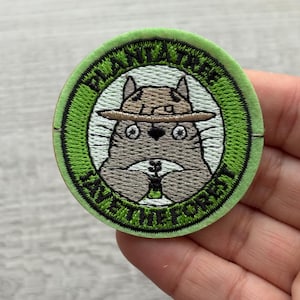 May include: Circular embroidered patch with a cartoon animal wearing a hat, and the text "PLANT A TREE SAVE THE FOREST" in green and white. The patch has a green border and is designed for application on clothing or accessories.