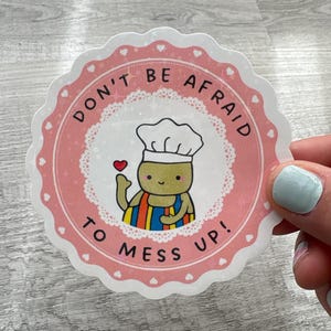 May include: A circular sticker with a pink scalloped border and the words "DON'T BE AFRAID TO MESS UP!". It features a cartoon chef holding a heart, wearing a white hat and a colourful apron.