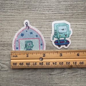 May include: Two stickers featuring a cartoon character. One sticker is a blue and white ornament shape with the character inside. The other sticker is the character on a skateboard. The stickers are approximately 1 to 2 inches in size.