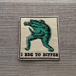 May include: A square embroidered patch with a cream-colored background and a black border. The design features a green frog holding a small knife, with the black embroidered text "I BEG TO DIFFER" positioned at the bottom of the patch.