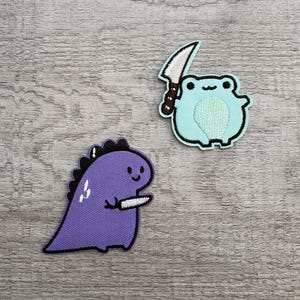 Puede incluir: Dos parches bordados: un dinosaurio morado y una rana azul claro, ambos sosteniendo cuchillos. El dinosaurio tiene una espina dorsal negra y la rana tiene una barriga verde claro. Ambos parches tienen contornos negros.