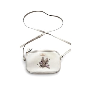 Może przedstawiać: Biała torba na ramię z dekoracyjnym wzorem ptaka. Torba posiada zapięcie na zamek błyskawiczny, długi regulowany pasek oraz logo Coach. Wzór ptaka jest ozdobiony cekinami i koralikami. Torba jest wykonana ze skóry.