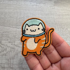 May include: Embroidered patch featuring a cartoon cat in an astronaut helmet. The cat is orange with black outlines and a white belly. The helmet is light blue with a black outline. The patch is likely for clothing or accessories.