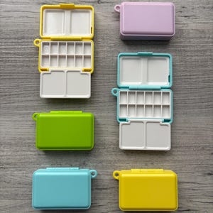 Könnte beinhalten: Eine Sammlung kleiner, rechteckiger Aquarellfarbpaletten in verschiedenen Farben, darunter Gelb, Lila, Grün und Blau. Jede Palette hat einen Klappdeckel und einen weißen Innenraum mit Farbnäpfen und Mischbereichen. Die Paletten liegen auf einer Holzoberfläche.