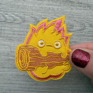 May include: Embroidered patch depicting a cartoon fire demon with yellow flames edged in pink, holding a brown log. The demon has white eyes and a smiling expression. The patch is designed for application on clothing or accessories.
