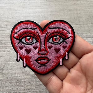 Op de afbeelding: Geborduurde hartvormige patch met een gezichtsontwerp. De patch heeft een rood-roze kleurenschema met een zwarte rand. Het gezicht heeft rode ogen, roze lippen en tranen. Kleine hartvormen zitten op de wangen.