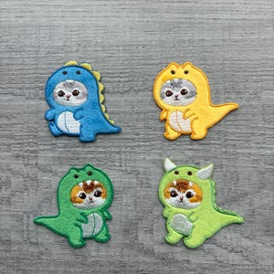 May include: Four embroidered patches featuring cats dressed as dinosaurs. The patches are in blue, yellow, and two shades of green. Each patch has a cat face with embroidered details and a dinosaur costume.