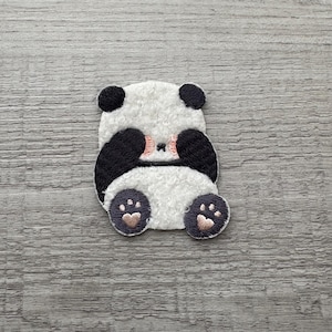 Op de afbeelding: Een geborduurde patch met een witte en zwarte pandabeer met roze wangen en zwarte ogen. De panda zit met zijn poten voor zijn gezicht.