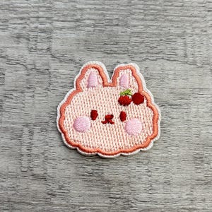 May include: Embroidered patch of a pink bunny face with a cherry bow. The patch has a white border and pink accents on the ears, cheeks, and nose. The cherries are red with green leaves.