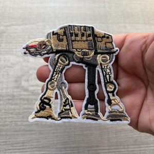 May include: Embroidered patch featuring an AT-AT walker from Star Wars. The patch has a tan, black, and gray color scheme with intricate detailing. The patch is approximately 3 inches tall and 3.5 inches wide.