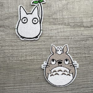 Könnte beinhalten: Zwei aufgestickte Aufnäher mit Figuren aus einem beliebten Zeichentrickfilm. Ein Aufnäher ist ein weißes Wesen mit großen Augen und einem grünen Blatt. Der andere ist ein graues Wesen mit einem kleineren Wesen auf dem Kopf.