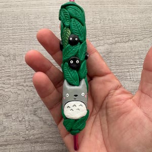 May include: A decorative pen featuring a gray Totoro character with a white belly, surrounded by green leaves and black soot sprites. The pen has a pink metal tip and is approximately 6 inches long.