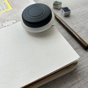 May include: A white watercolor ink bottle with a black cap sits atop a blank, textured watercolor paper pad. A wooden paintbrush and two watercolor paint blocks are also visible, suggesting art supplies for painting.