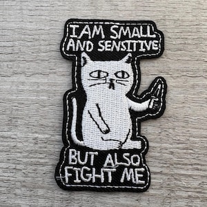 Könnte beinhalten: Gestickter Aufnäher mit einer weißen Katze, die ein Messer hält, mit schwarzen Rändern. Der Text auf dem Aufnäher lautet: "I AM SMALL AND SENSITIVE BUT ALSO FIGHT ME".