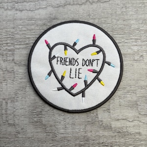Könnte beinhalten: Gestickter Aufnäher mit weißem Hintergrund und schwarzem Rand. Das Design zeigt eine schwarze Herzform mit den Worten "FRIENDS DON'T LIE" in der Mitte. Das Herz ist von bunten Lichterketten in Pink, Gelb und Blau umgeben.