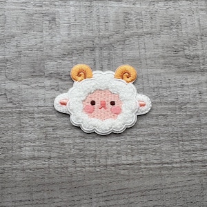 Op de afbeelding: Geborduurde patch met een cartoon schaap met een pluizig wit lichaam, roze gezicht en gouden hoorns. Het schaap heeft een lachende uitdrukking en is omlijnd met een witte rand. De patch is ontworpen voor kleding of accessoires.