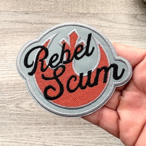 May include: Embroidered patch with the words "Rebel Scum" in black script. The patch features a red and white emblem on a gray background, with a gray border. The patch is round with a unique shape.