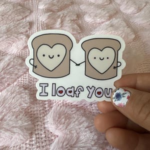 Könnte beinhalten: Ein weißer Aufkleber mit zwei cartoonartigen Brotscheiben mit Herzen in der Mitte. Die Brotscheiben halten sich an den Händen und haben fröhliche Gesichter. Der Text "I loaf you" befindet sich unter den Brotscheiben mit einem kleinen Herzen anstelle des "o".