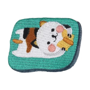 May include: Embroidered patch featuring a cartoon cat lying down, hugging a small yellow object. The cat has white fur with brown and black markings, pink cheeks, and a black nose and whiskers. The patch is on a teal green background with a dark blue border.