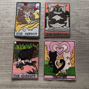 May include: Four rectangular embroidered patches featuring cat-themed tarot card designs. The patches depict The Hermit with a brown cat, The Emperor with a black and white cat on a throne, The Chariot with a cat on a skateboard, and The Lovers with two cats forming a heart shape with their tails.