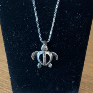 May include: A silver-toned necklace featuring a small, stylized sea turtle pendant with a cut-out shell design. The pendant hangs from a delicate, thin metal chain, showcasing a polished finish that reflects light across its smooth, curved surface.