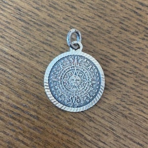 May include: A circular silver-toned pendant featuring an intricate embossed design of the Aztec sun stone calendar. The pendant has a textured, grooved border and includes a small silver jump ring at the top for attachment to a chain or cord.