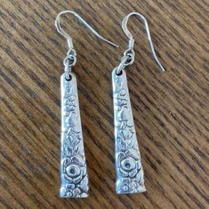 May include: A pair of silver-toned dangle earrings featuring a tapered, flat rectangular shape with an embossed floral pattern. Each earring is suspended from a simple curved metal ear wire, showcasing a detailed, textured design on the front surface.