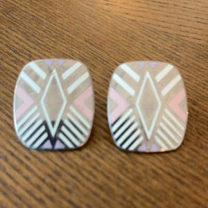 May include: A pair of square-shaped earrings with rounded corners, featuring a geometric pattern in shades of white, soft pink, and light purple, accented by thin metallic lines that form a central diamond shape and radiating stripes.
