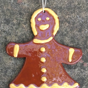 May include: A ceramic gingerbread man ornament with a glossy brown finish and yellow accents. The ornament has a smiling face, three yellow buttons, and a scalloped edge. A white string is attached for hanging.