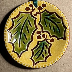 May include: A round ceramic ornament featuring a yellow background with a border of small raised dots. The center displays three green holly leaves with dark brown outlines and clusters of small brown berries. A dark green ribbon is attached at the top.