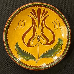 May include: A round, hand-painted ceramic plate with a yellow background and a central floral design in brown and green. The plate's rim is brown, with a green and brown scalloped border. The floral design features a stylized tulip.