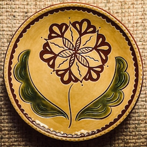 May include: A round, hand-painted ceramic plate with a floral design. The plate features a yellow background with a large brown flower in the center, green leaves, and a scalloped brown border. The plate is likely 10 inches in diameter.