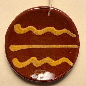 May include: A round, brown ceramic ornament with a glossy finish. The ornament features three yellow, wavy and straight lines. A white string is attached for hanging. The ornament is likely handmade.