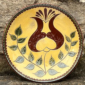 May include: A yellow ceramic plate with a brown and green floral design. The plate has a scalloped edge and is decorated with a stylized flower with leaves.