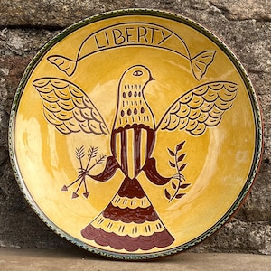 May include: A decorative ceramic plate with a yellow background and a brown and yellow bird design. The bird holds arrows and a branch, with the word "LIBERTY" on a banner above. The plate has a green and red rim.