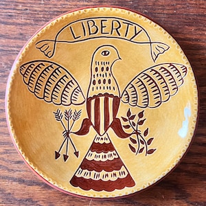 May include: A decorative ceramic plate with a yellow background and a brown eagle design. The word "LIBERTY" is written across the top. The plate has a red rim and is likely for display.